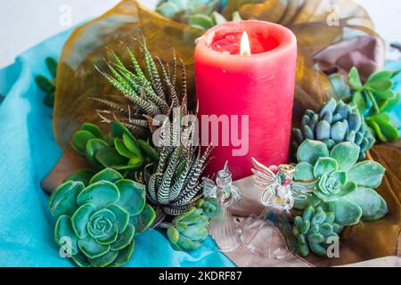 Sommerweihnachtsmittelpunkt einer rot angezündeten Kerze, umgeben von Sukkulenten, glänzenden Stoffen und Glaswinkelornamenten, Stockfoto