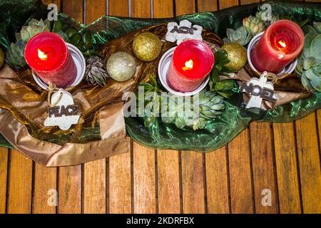 Rote Kerzen, umgeben von bronzefarbenen und grünen Stoffen, Sukkulenten und rustikalen Weihnachtsdekorationen aus Holz, bilden eine sommerliche Weihnachtsdekoration Stockfoto