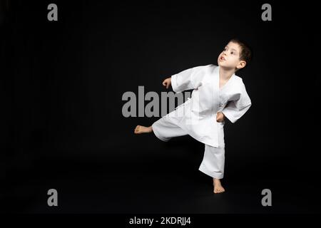 Ein kleiner Junge in einem Kimono übt Karate auf schwarzem Hintergrund und tritt nach vorne. Stockfoto