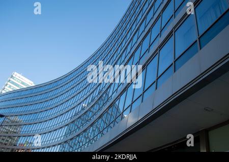 Blick auf das geschwungene Bürogebäude mit Spiegelung anderer Gebäude gegen den blauen Himmel Stockfoto