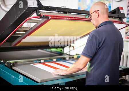 In der Druckerei arbeitet ein erfahrener Techniker an einer Siebdruckmaschine. Produktionsarbeit. Überprüfen Sie die Druckqualität. Stockfoto