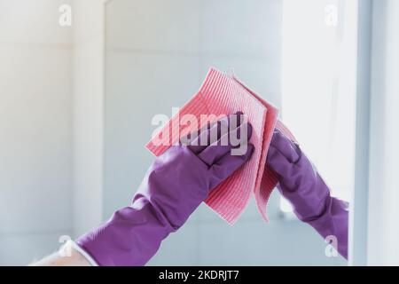 Beschnittene Ansicht der Haushälterin Frau Hand in lila Gummihandschuhe Polieren Spiegel mit rosa Tuch Putzer beim Reinigen Badezimmer zu Hause. Reinigungs- und Reinigungsservice. Sauberes Haus, Sauberkeit Stockfoto