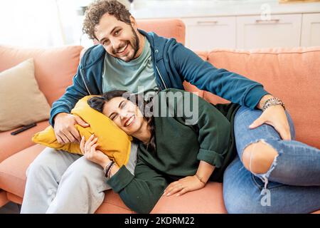 Glücklich, frisch verheiratet, sitzend auf der Couch und schauend auf die Kamera - ein paar junge Leute lächelnd, die sich auf dem Sofa in ihrem neuen Haus ausruhen - die Leute sind erhaben Stockfoto