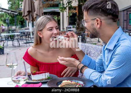 Glückliches romantisches Paar füttert sich nach dem Abendessen oder Mittagessen mit Desserts in einem wunderschönen Restaurant im Freien. Stockfoto