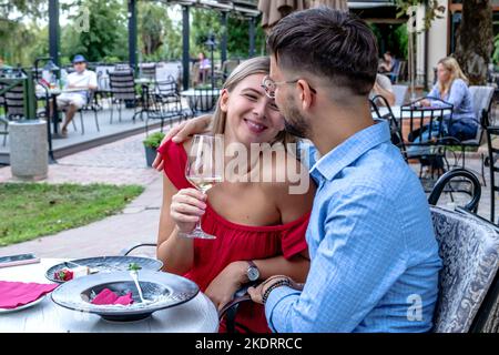 Glückliches romantisches Paar, das sich mit Wein zum Abendessen in einem wunderschönen Restaurant im Freien umarmt und anspricht. Lebensstil, Liebe und Beziehungen. Stockfoto