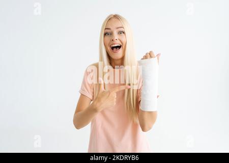 Studioportrait einer lachenden jungen Frau, die mit Zeigefingern auf einen gebrochenen Arm zeigt, eingewickelt in einen Gipsverband und auf die Kamera schaut, die auf Weiß steht Stockfoto