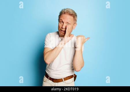 Porträt eines reifen Mannes in weißem T-Shirt, der mit Handfläche zu Mund posiert und Geheimnisse auf blauem Studiohintergrund flüstert Stockfoto