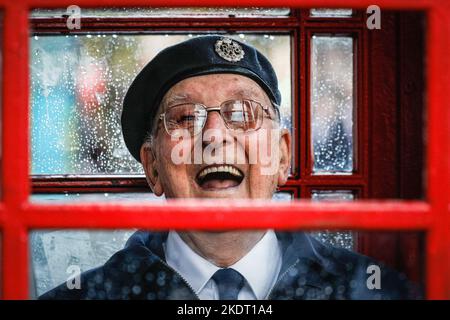 London, Großbritannien. 08.. November 2022. Ein britischer Militärveteran lächelt, als er sich in einer roten Londoner Telefonbox in Whitehall in der Nähe des Cenotaph vor dem starken Regen schützt. Kredit: Imageplotter/Alamy Live Nachrichten Stockfoto