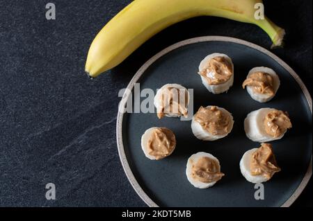 Gesunder Snack mit Bananenscheiben und knackiger Erdnussbutter auf dunklem Hintergrund mit Platz zum Kopieren Stockfoto