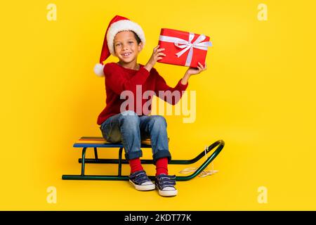 In voller Länge Foto von niedlichen kleinen Schüler Junge sitzen Schlitten halten Geschenk-Box Bogen gekleidet stilvolle rot gestrickt aussehen isoliert auf gelbem Hintergrund Stockfoto