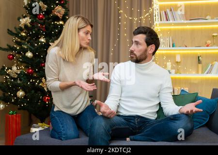 Familienkonflikt während des Urlaubs. Ein junges Paar, eine Familie, ein Mann und eine Frau streiten sich zu Weihnachten. Sie sitzen zu Hause auf dem Sofa in der Nähe des Weihnachtsbaums und brüllen sich gegenseitig an. Stockfoto