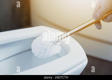 Nahaufnahme der Hände der Hausfrau in Handschuhen, die Toilettenschüssel mit Schrubber im Badezimmer oder in der öffentlichen Toilette reinigen. Desinfektion, Hygiene, Reinigungsdienstkonzept. Weibliche Wäsche, desinfizierender Toilettensitz. Stockfoto