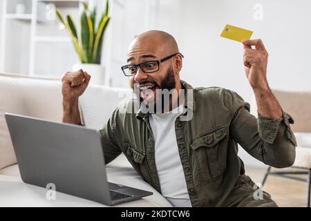 Online-Shopping. Fröhlicher lateinischer Mann mit Laptop und Kreditkarte, gestikuling ja, während erfolgreiche Zahlung Stockfoto