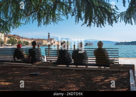 Ältere Menschen im Sommer, Rückansicht einer Gruppe älterer Menschen, die auf einer Bank sitzen und die malerische Stadt am Gardasee in Salo, Lombardei, Italien, betrachten Stockfoto