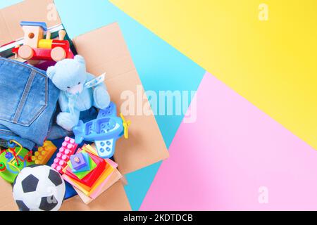 Spendenbox mit Kinderspielzeug, Büchern, Kleidung für wohltätige Zwecke auf buntem Gelb, Rosa, hellblauem Hintergrund. Draufsicht Stockfoto
