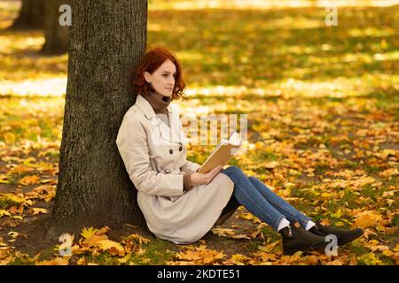 Serious smart kaukasische tausendjährige Frau mit roten Haaren in Regenmantel liest Buch und denkt, sitzt in der Nähe von Baum Stockfoto