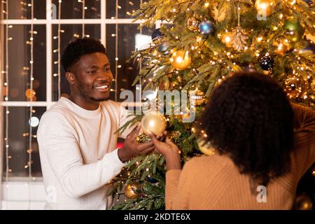 Fröhliche junge schwarze Paar Dekoration Weihnachtsbaum mit Girlanden, Spielzeug und Dekor in gemütlichen Wohnzimmer Stockfoto