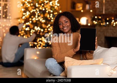 Fröhliche tausendjährige schwarze Frau zeigen Tablette mit leerem Bildschirm, Mann Dekoration Baum im Zimmer Innenraum Stockfoto