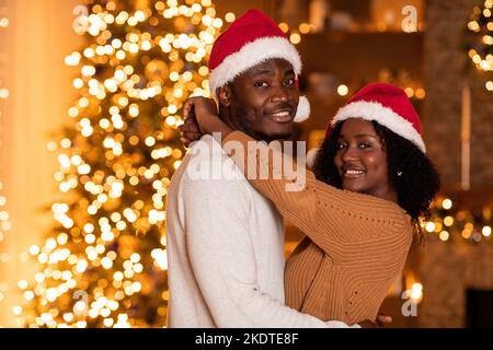 Fröhliche junge afroamerikanische Frau umarmt Mann in Weihnachtsmannhut in der Nähe von Weihnachtsbaum mit Lichtern Stockfoto