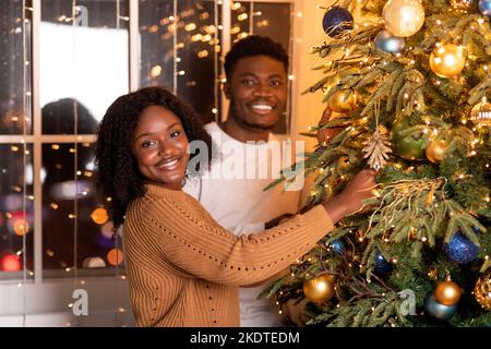 Glückliche tausendjährige afroamerikanische Frau und Mann schmücken Weihnachtsbaum mit Girlanden, Lichter Stockfoto