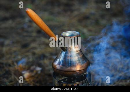 Kaffeeprozess am Lagerfeuer. Nahaufnahme Kaffee in türkischem Cezve auf Campingkocher. Tragbarer Herd, der Holzspäne verbrannte. Stockfoto