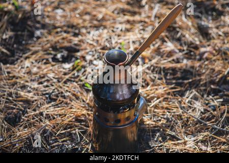 Kaffeeprozess am Lagerfeuer. Nahaufnahme Kaffee in türkischem Cezve auf Campingkocher. Tragbarer Herd, der Holzspäne verbrannte. Stockfoto