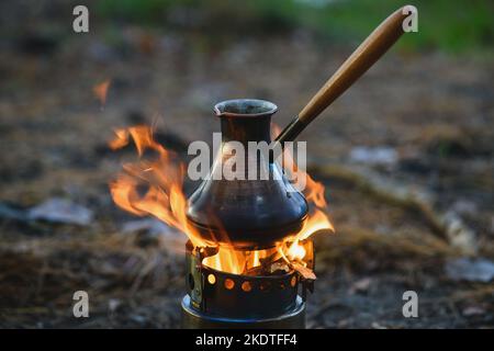 Kaffeeprozess am Lagerfeuer. Nahaufnahme Kaffee in türkischem Cezve auf Campingkocher. Tragbarer Herd, der Holzspäne verbrannte. Stockfoto