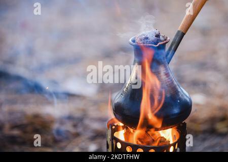 Kaffeeprozess am Lagerfeuer. Nahaufnahme Kaffee in türkischem Cezve auf Campingkocher. Tragbarer Herd, der Holzspäne verbrannte. Stockfoto