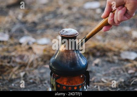 Kaffeeprozess am Lagerfeuer. Nahaufnahme Kaffee in türkischem Cezve auf Campingkocher. Tragbarer Herd, der Holzspäne verbrannte. Stockfoto