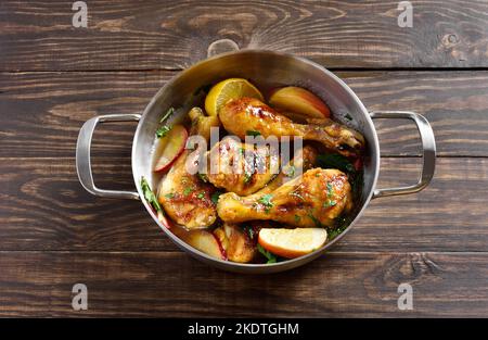 Hähnchendrumsticks, gebacken mit Äpfeln und Kräutern in einem Brühtopf auf Holzboden. Nahaufnahme Stockfoto