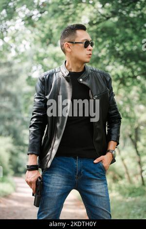 Ein Mann in einer dunklen Brille und einer schwarzen Lederjacke hält eine Waffe in den Händen Stockfoto