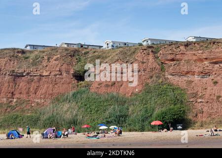 Touristen am Sandy Bay Beach mit Ferienpark statische Wohnwagen auf den roten Sandsteinklippen oben, Exmouth, Devon, Großbritannien, August. Stockfoto