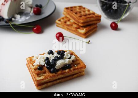 Das Frühstück ist gesund mit Hüttenkäse und Beeren. Belgische weiche Waffeln mit Hüttenkäse-Blaubeeren und Kirschen auf weißem Tischhintergrund. H Stockfoto