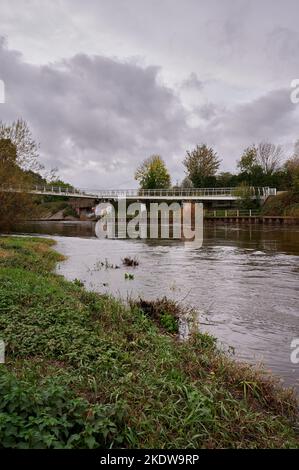 07 Nov 2022 - Hereford UK: Fußgängerbrücke mit Graffiti über dem geschwollenen Fluss unter wolkendem Himmel vom Flussufer Stockfoto