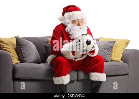 Überraschte den weihnachtsmann mit einem Fußball und sitzt auf einem Sofa isoliert auf weißem Hintergrund Stockfoto