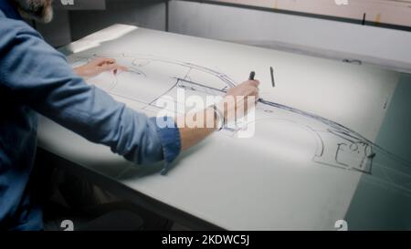 Senior Automobil-Designer Zeichnung Karosserie Skizze mit Marker am Lichttisch, Entwicklung futuristischer Elektroauto Zukunft Design. Er arbeitet im Entwicklungsstudio für modernes Fahrzeugdesign. Stockfoto