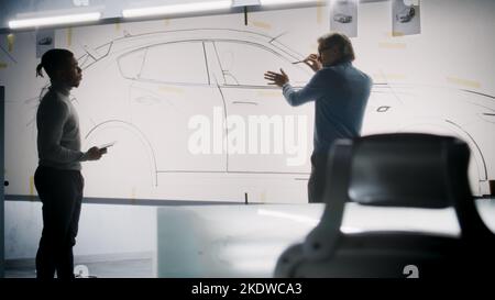 Zwei verschiedene männliche Automobildesigner diskutieren und entwickeln futuristisches Elektroauto-Design. Eine Zeichnung Skizzen auf Whiteboards, andere halten Tablet-Computer. Auto-Design-Entwicklungsstudio. Stockfoto