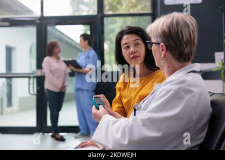 Leitender Arzt, der einen asiatischen Patienten mit einem Glucometer-Instrument konsultiert und den Insulin- und Glukosespiegel aus der Blutprobe misst. Arzt und Frau, die Diabetes-Kontrolle beim Termin für den Kontrollbesuch machen. Stockfoto