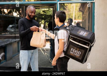 Food Delivery Service Kurier liefert Mittagessen ins Büro, Frau gibt Kunden Lunch Papiertüte. Lächelnder afroamerikanischer Gast nimmt Essen zum Mitnehmen im Freien, mittlere Aufnahme Stockfoto