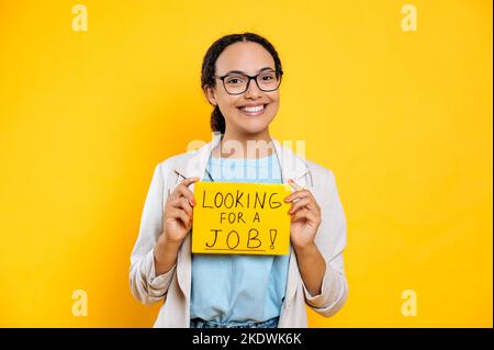 Aufgeregt positive erfolgreiche brasilianische oder hispanische junge Frau, kreative Spezialistin auf der Suche nach einem Job, steht auf isoliertem orangefarbenem Hintergrund, zeigt ein Schild auf der Suche nach einem Job, schaut lächelnd in die Kamera Stockfoto