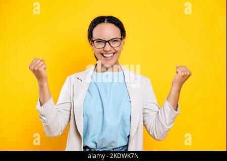 Gewinnen, Erfolg. Überglücklich gemischter Rennbusiness Frau mit Brille, freut sich über gute Nachrichten, Sieg, Deal, gestikulieren mit Fäusten, steht auf isoliertem orangefarbenem Hintergrund, schaut auf die Kamera und lächelt fröhlich Stockfoto