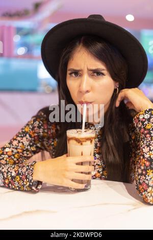 Wütend auf die Kamera schauen und einen Drink mit Stroh, Gesichtsausdruck und Getränke, junge latinerin mit langen Haaren und modischem Hut Stockfoto