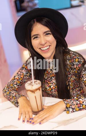 Lächelnde junge lateinerin mit langen Haaren und Hut, Lifestyle und Schönheit mit leckerem Getränk, Dessert Stockfoto