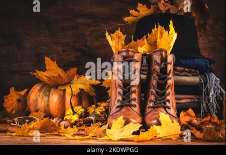 Herbsthintergrund mit braunen Lederstiefeln, herbstlicher warmer Kleidung, Halstüchern und Filzhut, Kürbissen, Ahorn- und Eichenblättern auf rustikalem Holzhintergrund. Land Stockfoto