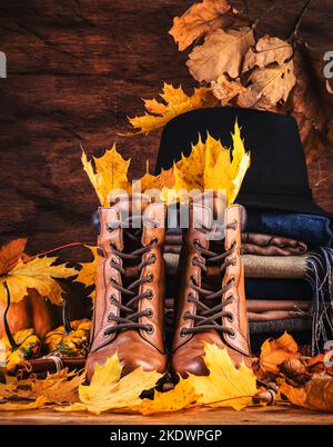 Herbsthintergrund mit braunen Lederstiefeln, herbstlicher warmer Kleidung, Halstüchern und Filzhut, Kürbissen, Ahorn- und Eichenblättern auf rustikalem Holzhintergrund. Land Stockfoto