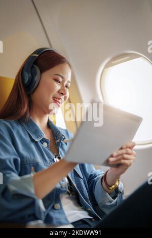 Attraktives Porträt einer asiatischen Frau, die in der Economy-Klasse auf einem Fenstersitz sitzt und während eines Flugzeugs Instrumentalmusik hört Stockfoto
