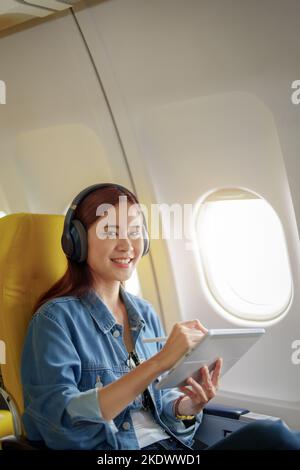 Attraktives Porträt einer asiatischen Frau, die in der Economy-Klasse auf einem Fenstersitz sitzt und während eines Flugzeugs Instrumentalmusik hört Stockfoto