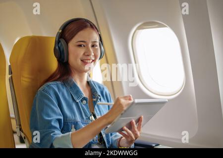 Attraktives Porträt einer asiatischen Frau, die in der Economy-Klasse auf einem Fenstersitz sitzt und während eines Flugzeugs Instrumentalmusik hört Stockfoto