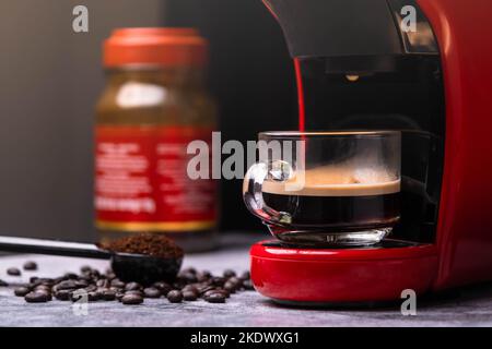 Heißer Kaffee fließt in eine Tasse mit einer Espressomaschine, Nahaufnahme. Moderne Espressomaschine mit gemahlenem Kaffeesatz in einem Löffel und verschütteten Kaffeebohnen Stockfoto