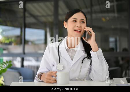 Attraktive und professionelle junge asiatische Ärztin in weißem Kleid, die am Telefon mit jemandem an ihrem Schreibtisch telefoniert. Stockfoto